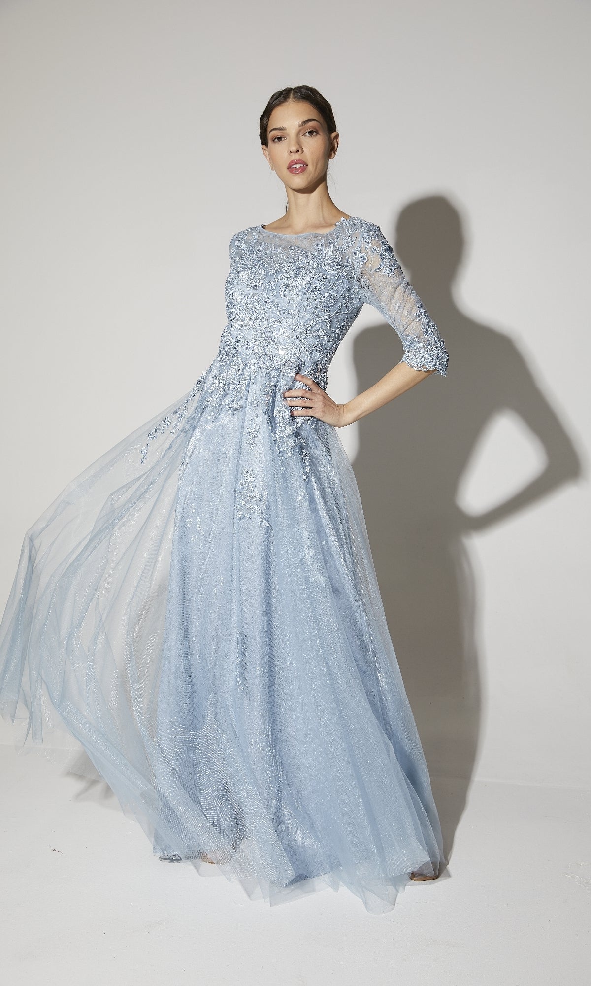 A woman models the Eureka 9660 Flowy Long Sleeve Long Formal Dress in light blue lace, featuring a three-quarter sleeve bodice, standing against a plain white background.